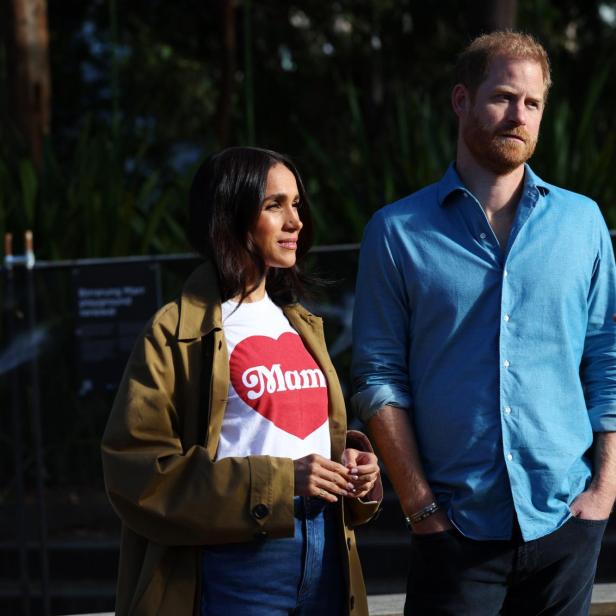 Meghan Markle und Prinz Harry stehen im Freien und unterhalten sich mit einer Person, die nur teilweise im Vordergrund zu sehen ist.