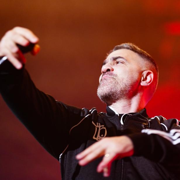 Bushido mit Bart und schwarzer Trainingsjacke hält ein Mikrofon und gestikuliert energisch vor rotem Hintergrund.