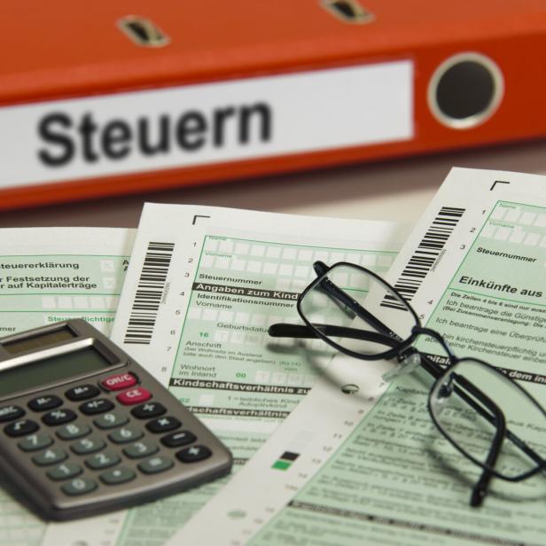 Ein Taschenrechner und eine Brille liegen auf ausgefüllten Steuerformularen vor einem roten Ordner mit der Aufschrift „Steuern“.