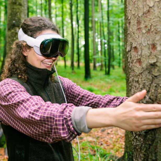 Eine Frau mit VR-Brille umarmt lachend einen Baum im Wald.