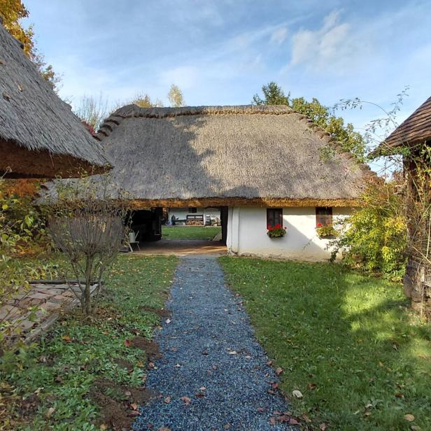 Ein schmaler Weg führt zu einem weißen Haus mit Strohdach, umgeben von alten Holzhäusern und herbstlicher Vegetation.