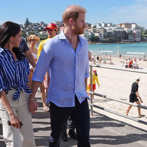 Harry und Meghan spazieren Hand in Hand am Bondi Beach entlang, im Hintergrund das Meer und Badegäste.