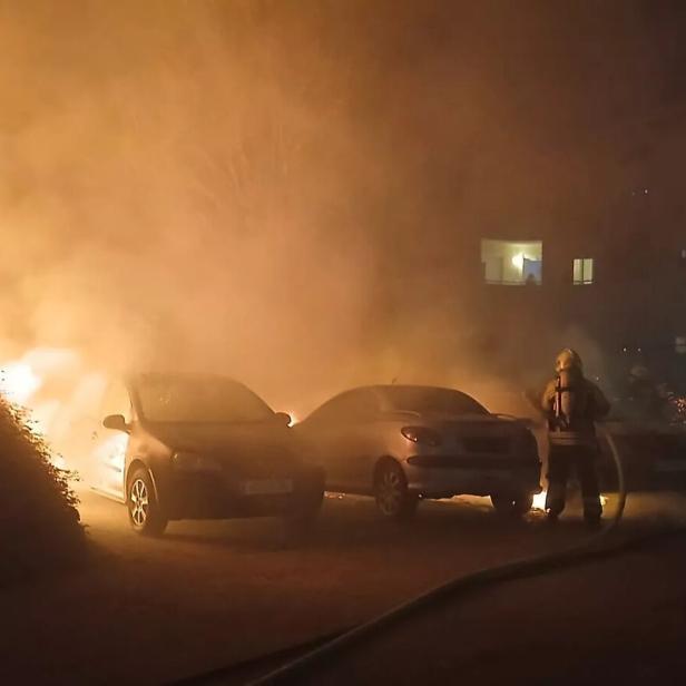 Starke Flammen und dichter Rauch umgeben mehrere Autos, während Feuerwehrleute versuchen, das Feuer zu löschen.
