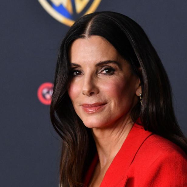 Sandra Bullock trägt einen roten Blazer vor einem dunklen Hintergrund mit dem Warner Bros. Logo.