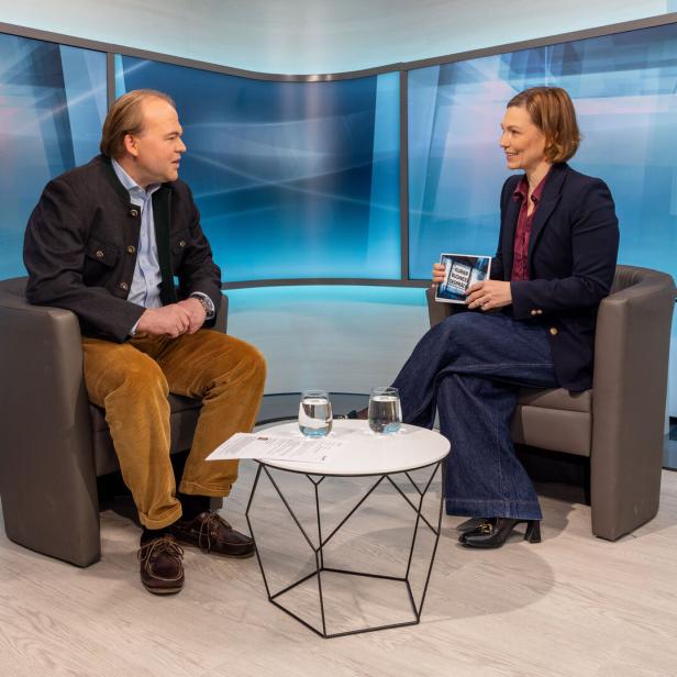 Zwei Personen sitzen sich in einem modernen Studio gegenüber, zwischen ihnen steht ein kleiner Tisch mit zwei Gläsern Wasser.