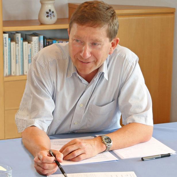 Mann sitzt an einem Tisch mit Unterlagen und Glas Wasser, Bücherregal im Hintergrund.