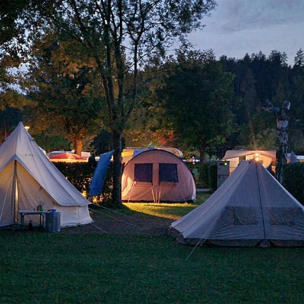 Zelte und Wohnwagen stehen abends beleuchtet auf einer grünen Wiese zwischen Bäumen.