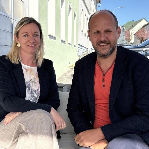 Zwei Personen sitzen nebeneinander auf einem Sofa im Freien und lächeln in die Kamera.