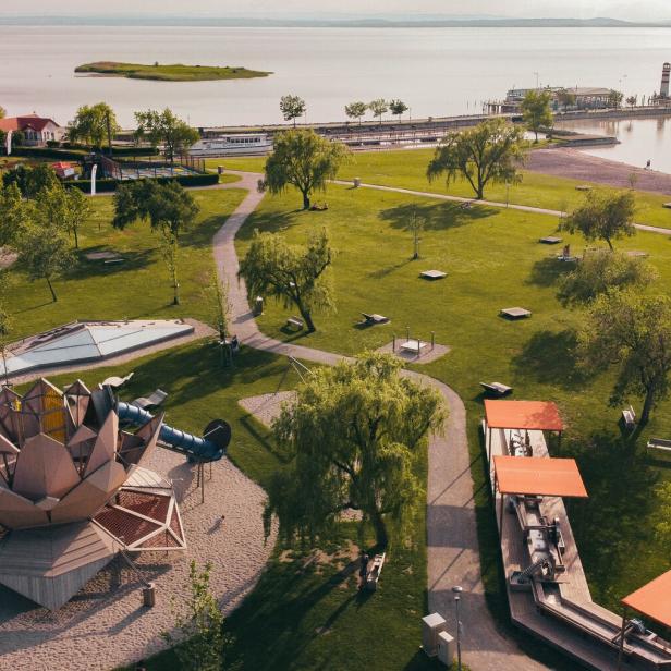 Großer grüner Park mit Spielplatz, Wegen, Bäumen und Sitzbereichen am Ufer eines ruhigen Sees mit Steg und Leuchtturm.