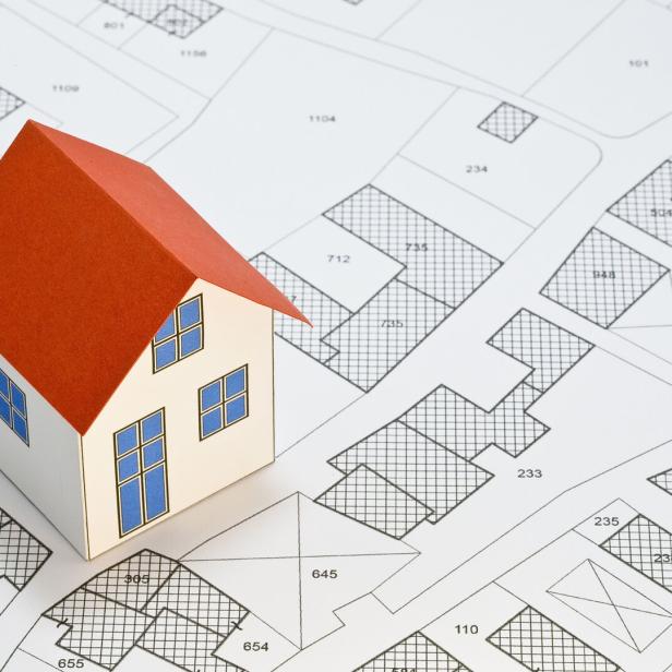 Ein Modellhaus mit rotem Dach steht auf einem detaillierten Stadtplan mit Grundstücksparzellen und Hausnummern.