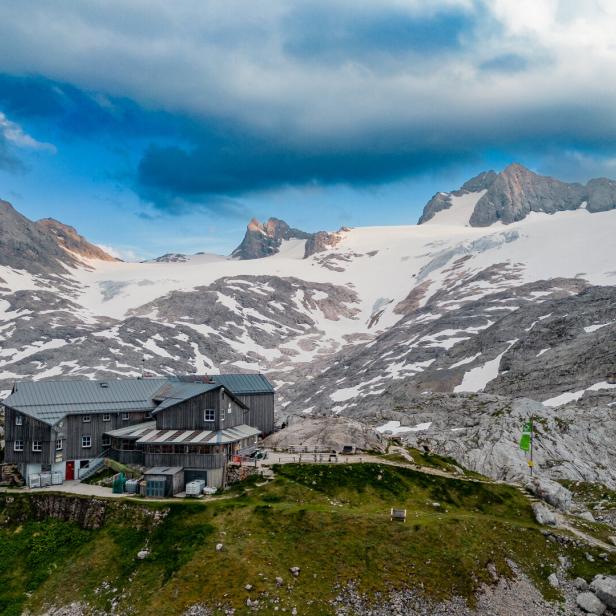 Berghütte mit mehreren Gebäuden auf grünem Hang, umgeben von schneebedeckten Felsen und hohen Bergen unter bewölktem Himmel.