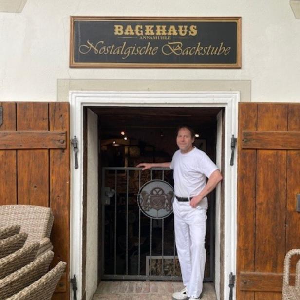 Ein Mann in weißer Kleidung steht in der Tür einer traditionellen Bäckerei mit Holzläden und nostalgischem Schild.
