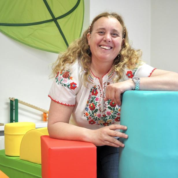 Eine lachende Frau mit lockigem Haar lehnt an bunten Schaumstoffblöcken in einem freundlich eingerichteten Raum.