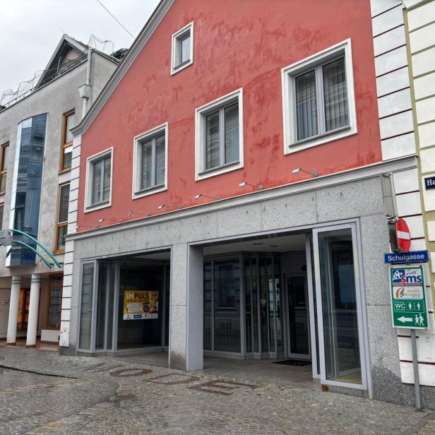Mehrstöckiges Gebäude mit roter Fassade, mehreren Fenstern und einem offenen Eingang an einer gepflasterten Straße.