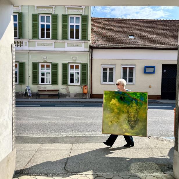 Eine Person mit heller Kurzhaarfrisur trägt ein großes, buntes Gemälde über eine sonnige Straße vor Altbauten.