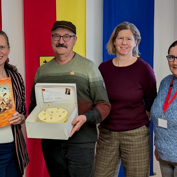 Vier Personen stehen lächelnd vor bunten Fahnen, eine Frau hält ein Buch, ein Mann eine Torte mit Karte.