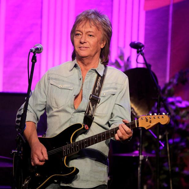 Chris Norman spielt Gitarre auf einer Bühne mit lila Hintergrundbeleuchtung.