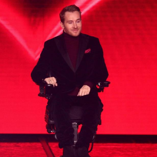 Samuel Koch im Rollstuhl vor leuchtend rotem Hintergrund, elegant gekleidet und mit leichtem Lächeln.