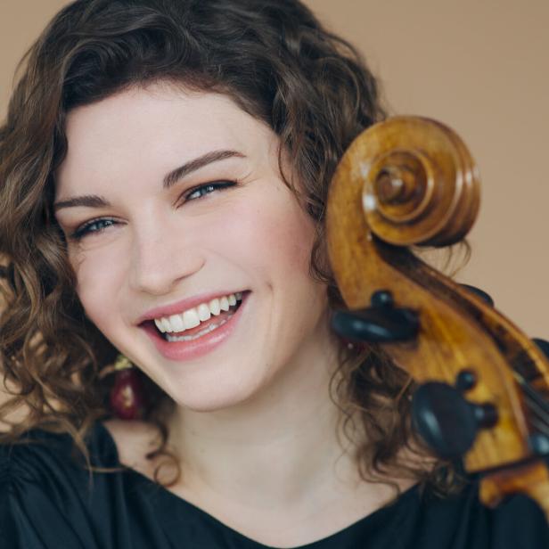 Junge Frau mit lockigen Haaren lächelt und hält ein Streichinstrument mit sichtbarem Wirbel neben ihrem Gesicht.
