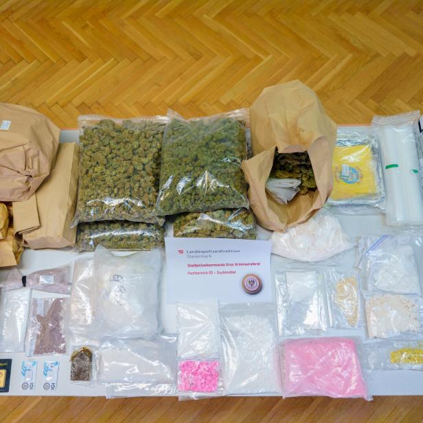 Auf einem Tisch liegen beschlagnahmte Drogen, darunter Cannabis, Pulver und Pillen, sowie eine Goldbarren und eine Waage.