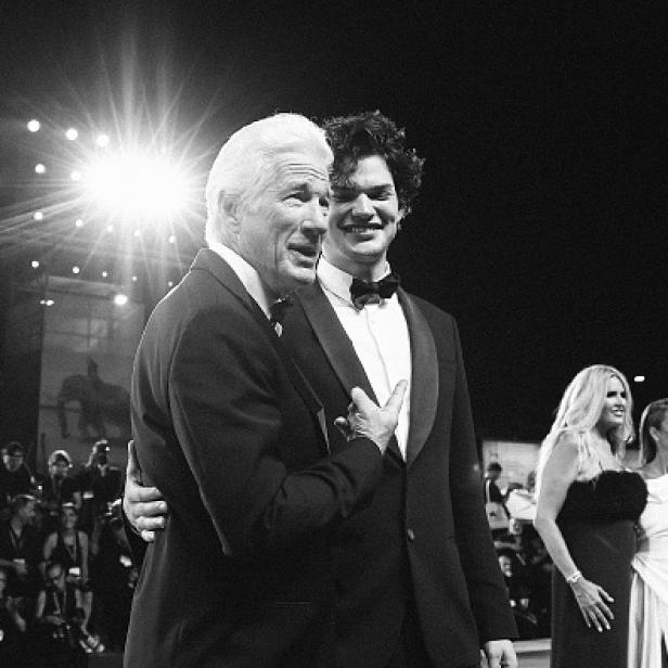 Richard Gere und sein Sohn Homer James Jigme Gere stehen gemeinsam im Anzug auf einem roten Teppich und lächeln, umgeben von weiteren Gästen.