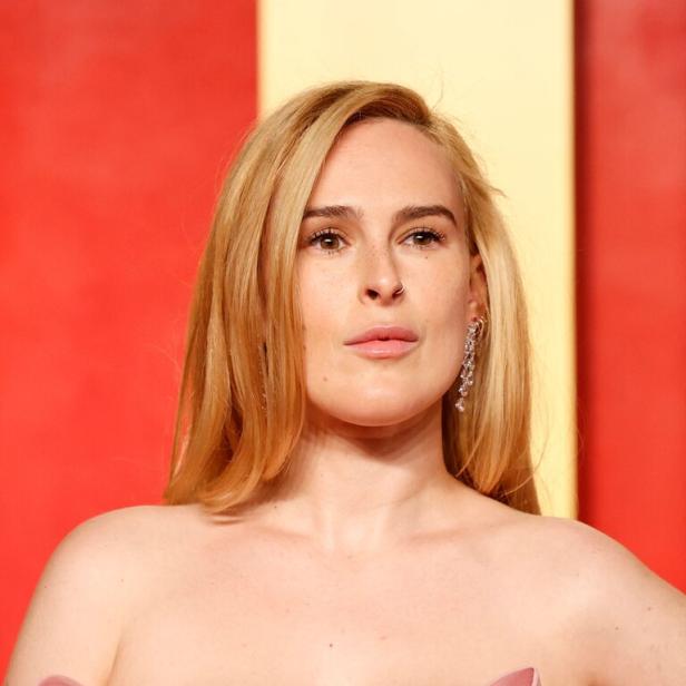 Rumer Willis mit blonden Haaren und einem rosa Kleid posiert vor einem roten Hintergrund.