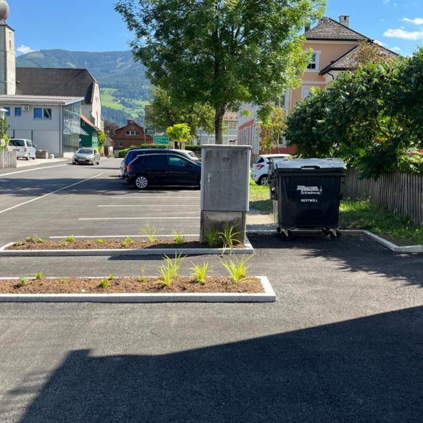 Frisch asphaltierter Parkplatz mit bepflanzten Grüninseln und umliegenden Gebäuden bei Sonnenschein.