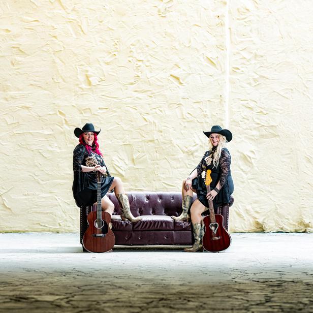 Zwei Frauen mit Cowboyhüten und Gitarren posieren auf einem Sofa vor heller Wand.