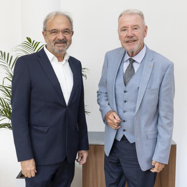 Zwei ältere Männer in Anzügen stehen nebeneinander vor einer weißen Wand mit einer Pflanze im Hintergrund.