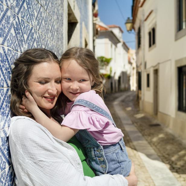Mutter und Tochter umarmen sich lachend in einer sonnigen Gasse mit blauen Fliesen.