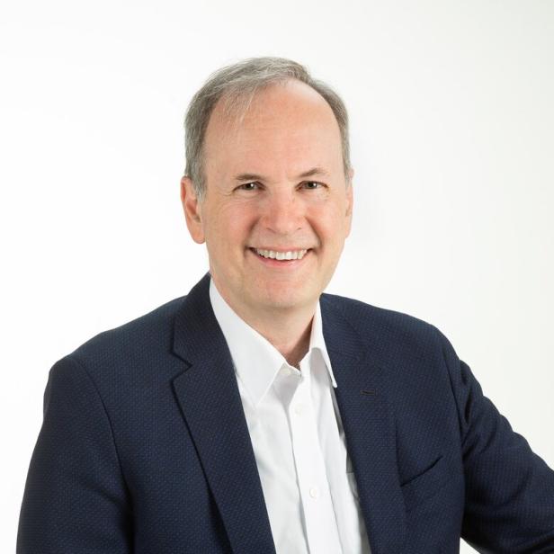 Mann mittleren Alters mit grauen Haaren trägt ein dunkelblaues Sakko und weißes Hemd und lächelt freundlich.