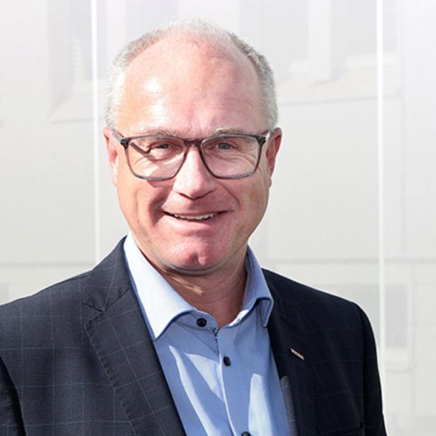 Ein älterer Mann mit Brille und Anzug lächelt vor einem hellen, unscharfen Hintergrund.