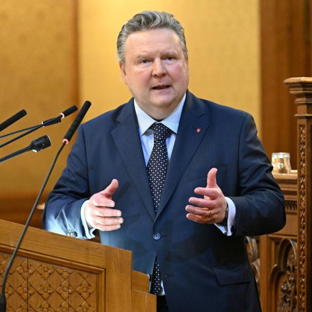 Ein Mann im Anzug spricht gestikulierend an einem Rednerpult mit mehreren Mikrofonen in einem holzverzierten Raum.