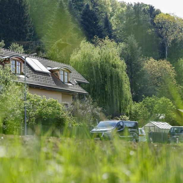 Ein Haus mit Dachfenstern steht vor einem bewaldeten Hügel, davor parken Autos.