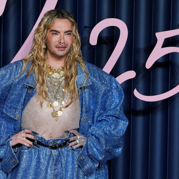 Bill Kaulitz mit blonden Locken, Nasenring und glitzernder Jeansjacke posiert vor blauem Hintergrund.