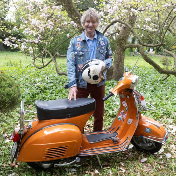 Ein älterer Mann steht im Garten neben einem orangefarbenen Motorroller und hält einen Helm in der Hand.