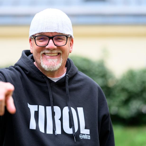 DJ Ötzi mit Brille und Mütze lächelt und zeigt auf den Betrachter, er trägt einen "Tirol"-Hoodie.