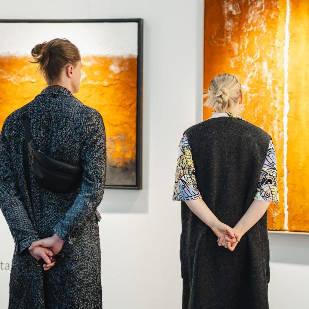 Zwei Personen betrachten abstrakte Gemälde in warmen Orangetönen an einer weißen Galeriewand.