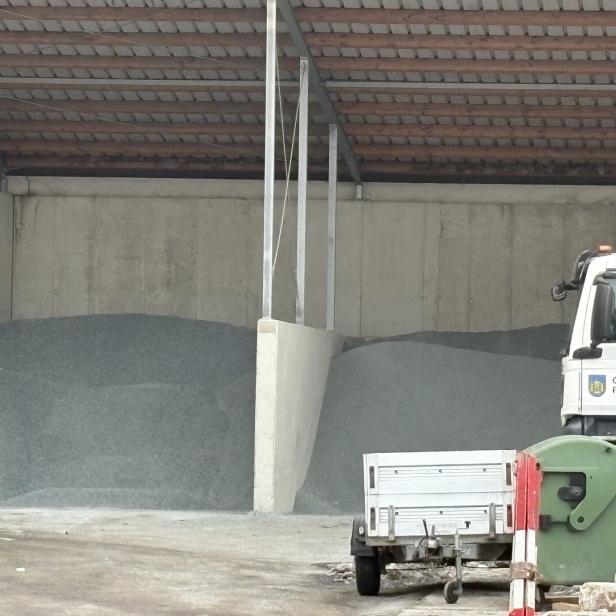Zwei Haufen Streusalz oder Splitt unter einem Dach, daneben ein Kommunalfahrzeug mit Anhänger und Müllcontainer.