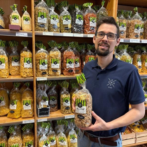 Ein Mann mit Brille steht vor Regalen voller verschiedener Nudelsorten und hält eine Packung Nudeln in der Hand.