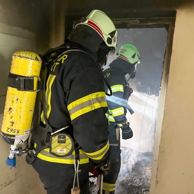 Zwei Feuerwehrleute mit Atemschutzgeräten betreten einen verrauchten Raum und leuchten mit einer Taschenlampe hinein.