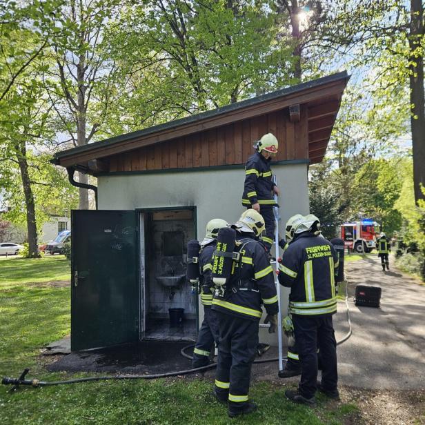 Feuerwehrleute untersuchen ein kleines Gebäude mit offener Tür und Brandschäden.