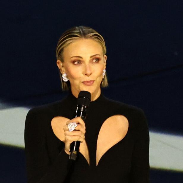 Charlize Theron mit blonden Haaren spricht in einem schwarzen Kleid mit Herz-Ausschnitt in ein Mikrofon.