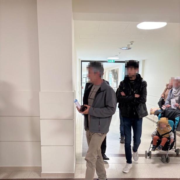 Mehrere Personen gehen durch einen hellen Flur, darunter ein Mann mit Dokumenten und ein Kind im Buggy.