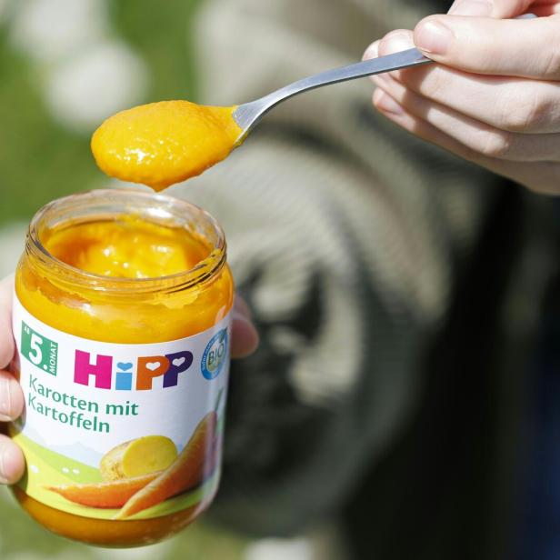 Ein Glas Hipp Karotten mit Kartoffeln Brei wird mit einem Löffel entnommen und von einer Hand gehalten.