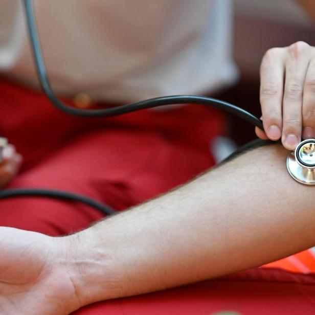 Eine Person misst mit einem Stethoskop und einer Blutdruckmanschette den Blutdruck am Arm einer anderen Person.