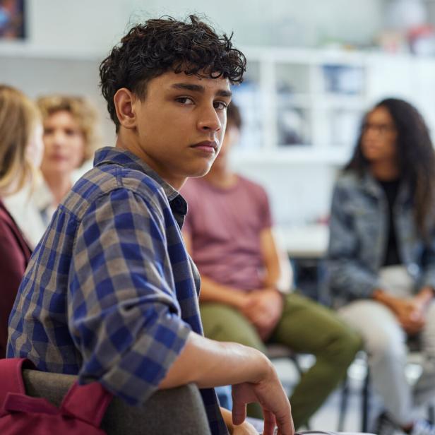 Ein Jugendlicher mit kariertem Hemd sitzt in einer Gruppe und blickt ernst zur Kamera, andere Jugendliche sind im Hintergrund.