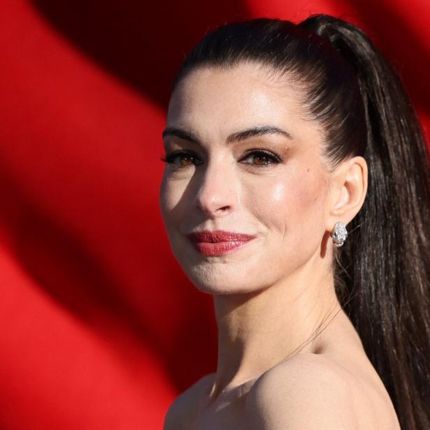 Anne Hathaway mit hochgesteckten, dunklen Haaren und Ohrringen vor rotem Hintergrund.