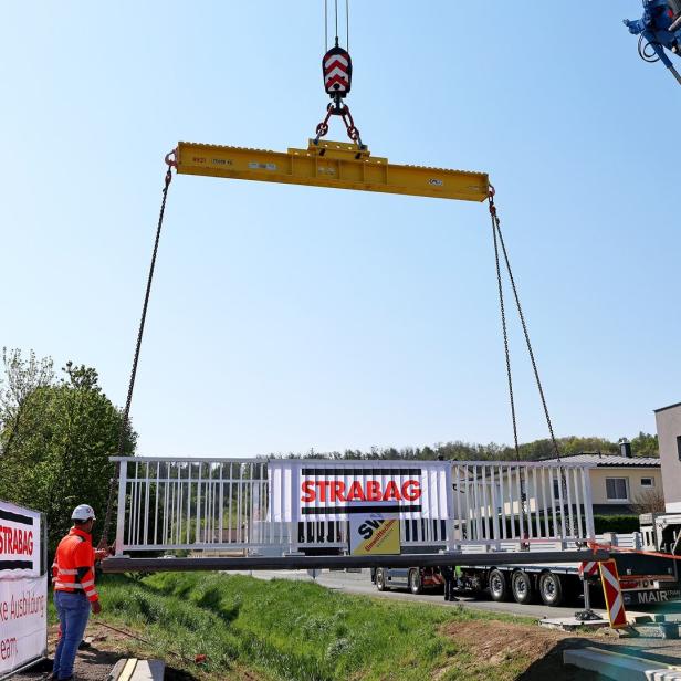 Ein Kran hebt eine weiße Brücke mit STRABAG-Logo, während zwei Bauarbeiter in Warnkleidung den Bereich sichern.