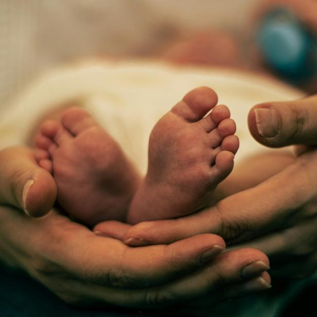 Erwachsene Hände halten sanft die Füße eines Babys, im Hintergrund ein Schnuller.